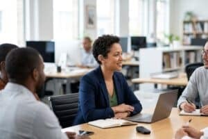 A group of professionals collaborating in a bright business meeting, featuring a woman wearing a discreet behind-the-ear hearing aid as she listens to a colleague.